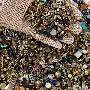 May include: A close-up of a collection of vintage jewellery components. The components include colourful rhinestones, beads, and metal findings in gold and silver tones. The jewellery components are held by a person wearing a white lace glove.
