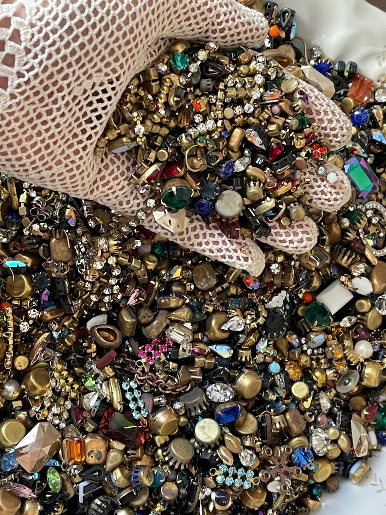 May include: A close-up of a collection of vintage jewellery components. The components include colourful rhinestones, beads, and metal findings in gold and silver tones. The jewellery components are held by a person wearing a white lace glove.