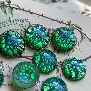 May include: A collection of six green and blue iridescent glass cabochons with gold foil accents. The cabochons are set in gold-toned bezels and have a speckled, textured surface.
