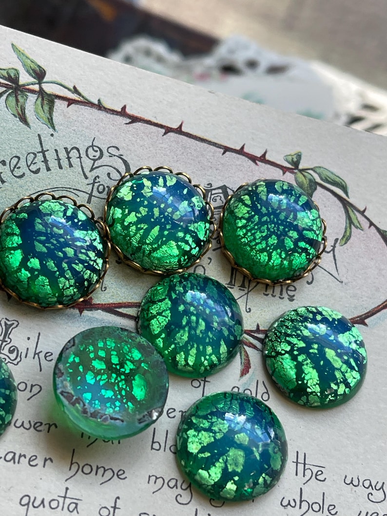 May include: A collection of six green and blue iridescent glass cabochons with gold foil accents. The cabochons are set in gold-toned bezels and have a speckled, textured surface.
