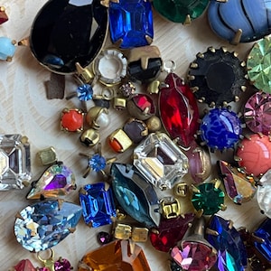 May include: A selection of colourful vintage rhinestone jewels in various shapes and sizes. The gemstones include black, blue, red, green, and pink, with gold-tone metal settings. The arrangement is on a light-coloured surface.