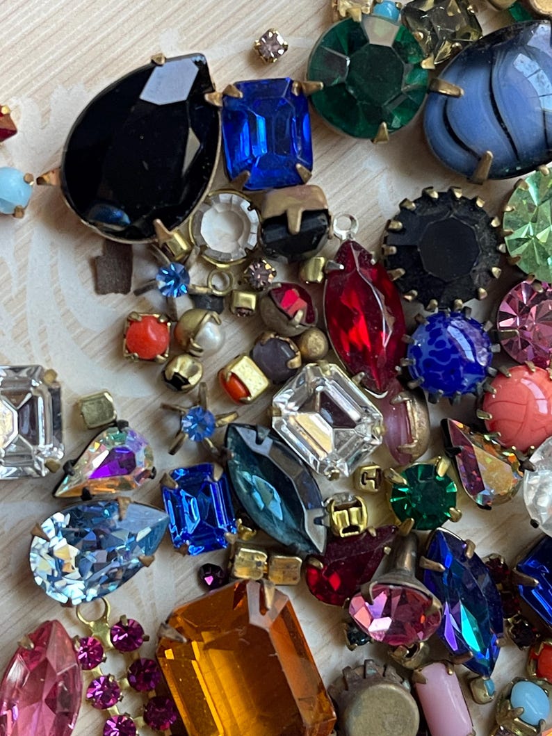 May include: A selection of colourful vintage rhinestone jewels in various shapes and sizes. The gemstones include black, blue, red, green, and pink, with gold-tone metal settings. The arrangement is on a light-coloured surface.