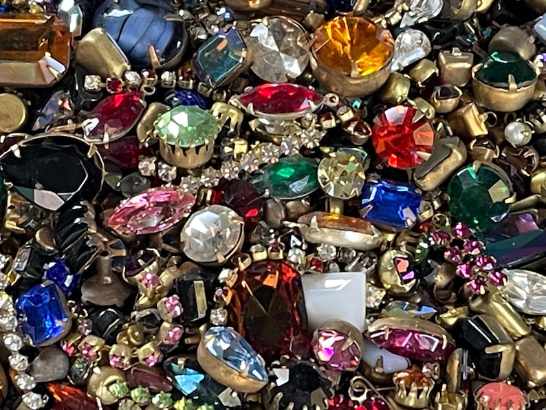May include: A close-up of a collection of colourful gemstones and rhinestones. The gems are in various shapes and sizes, including round, oval, and square. Colours include red, blue, green, yellow, and pink. The gems are set in gold-coloured metal.
