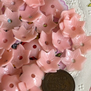 Peut inclure: Un ensemble de boutons en plastique rose en forme de fleur avec un trou au centre. Les boutons sont dispos&eacute;s en pile sur une surface blanche avec une pi&egrave;ce de monnaie vintage dans le coin inf&eacute;rieur droit.