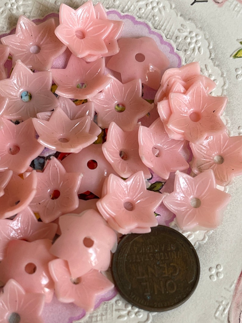 Peut inclure: Un ensemble de boutons en plastique rose en forme de fleur avec un trou au centre. Les boutons sont dispos&eacute;s en pile sur une surface blanche avec une pi&egrave;ce de monnaie vintage dans le coin inf&eacute;rieur droit.