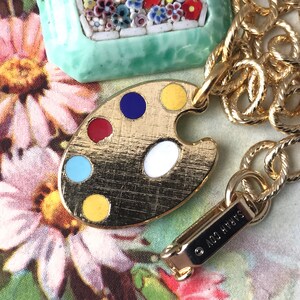 K&ouml;nnte beinhalten: Goldkette mit einem goldenen Farbpaletten-Anh&auml;nger mit roter, blauer, gelber, wei&szlig;er und t&uuml;rkisfarbener Emaille-Farbe. Der Anh&auml;nger ist an einem goldenen Karabinerhaken mit dem eingravierten Text "&copy; NICO HYATT" befestigt.