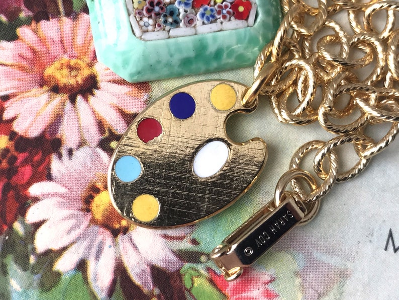 K&ouml;nnte beinhalten: Goldkette mit einem goldenen Farbpaletten-Anh&auml;nger mit roter, blauer, gelber, wei&szlig;er und t&uuml;rkisfarbener Emaille-Farbe. Der Anh&auml;nger ist an einem goldenen Karabinerhaken mit dem eingravierten Text "&copy; NICO HYATT" befestigt.
