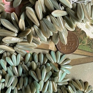 May include: A close-up of a pile of light green and brown glass beads. The beads are shaped like long, narrow leaves. There is a copper penny in the background.