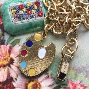 K&ouml;nnte beinhalten: Eine goldene Kettenkette mit einem gr&uuml;nen und wei&szlig;en Mosaik-Anh&auml;nger und einem goldenen Anh&auml;nger in Form einer Malpalette mit buntem Emaille-Anstrich. Die Kette hat einen Verschluss mit der Aufschrift "&copy; 1993 COV."