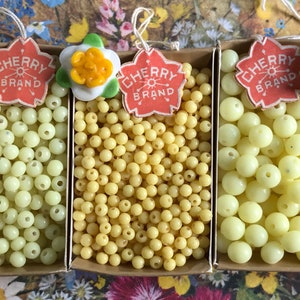 May include: Three boxes filled with pale yellow and cream colored glass beads. The boxes have red and white labels that say "Cherry Brand".