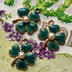 Puede incluir: Tres colgantes de tr&eacute;bol de cuatro hojas esmaltados en verde con bordes de metal dorado y un centro de perla de imitaci&oacute;n. Los colgantes est&aacute;n sobre un fondo decorativo con elementos florales verdes y morados.