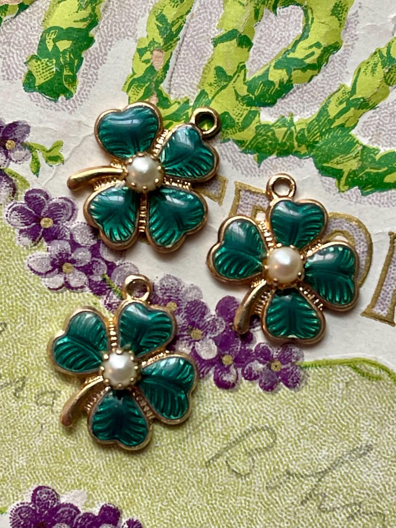 Puede incluir: Tres colgantes de tr&eacute;bol de cuatro hojas esmaltados en verde con bordes de metal dorado y un centro de perla de imitaci&oacute;n. Los colgantes est&aacute;n sobre un fondo decorativo con elementos florales verdes y morados.