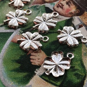 May include: Seven silver four-leaf clover charms with a loop for attaching to a necklace or bracelet. The charms are on a green and brown background.