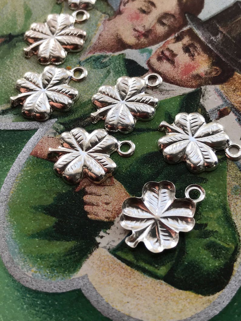 May include: Seven silver four-leaf clover charms with a loop for attaching to a necklace or bracelet. The charms are on a green and brown background.