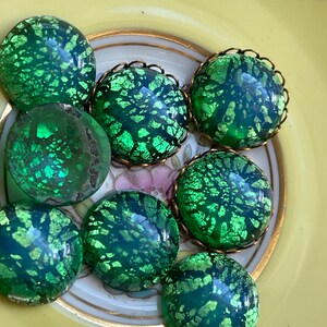 May include: A collection of eight green glass cabochons with a gold foil pattern. The cabochons are round and have a slightly iridescent finish.