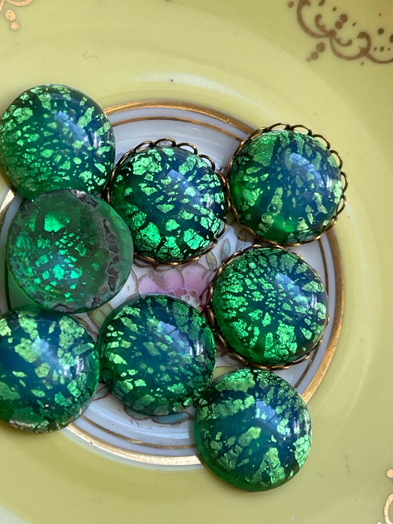 May include: A collection of eight green glass cabochons with a gold foil pattern. The cabochons are round and have a slightly iridescent finish.