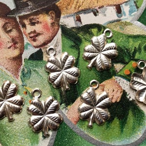 May include: Six silver four-leaf clover charms with a loop at the top for attaching to a necklace or bracelet. The charms are on a green and white background.