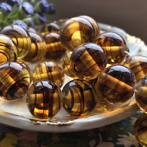 May include: A strand of amber glass beads with a swirling, striped pattern. The beads are round and have a translucent, golden brown color.