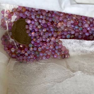 K&ouml;nnte beinhalten: Nahaufnahme eines wei&szlig;en Papierbeutels, der mit einer gro&szlig;en Menge kleiner, irisierender rosa und lila Perlen gef&uuml;llt ist. Eine einzelne M&uuml;nze ist am Boden des Beutels sichtbar.