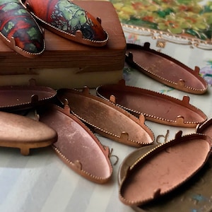 May include: A group of ten oval-shaped copper-colored metal settings with a beaded edge. The settings are designed to hold a cabochon stone or other decorative element.