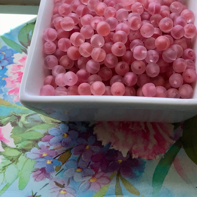 May include: A white bowl filled with pink frosted glass beads. The beads are round and have a slightly iridescent finish.