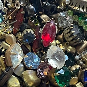 May include: A close-up of a collection of vintage jewellery components. The image features a variety of colourful rhinestones and gemstones in various shapes and sizes, including red, blue, green, and clear stones, set in gold-toned metal settings.
