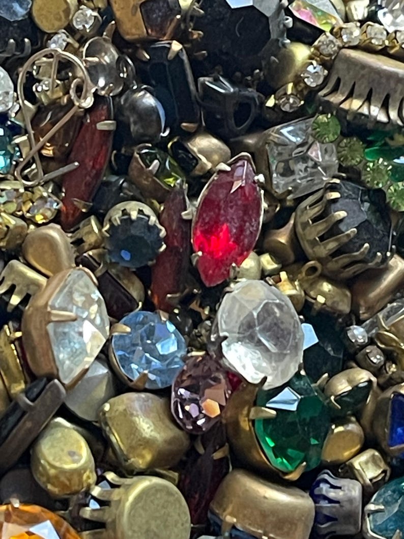May include: A close-up of a collection of vintage jewellery components. The image features a variety of colourful rhinestones and gemstones in various shapes and sizes, including red, blue, green, and clear stones, set in gold-toned metal settings.