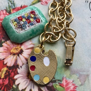 K&ouml;nnte beinhalten: Ein goldfarbenes Kettenarmband mit einem gr&uuml;nen Mosaik-Anh&auml;nger und einem goldenen Anh&auml;nger in Form einer K&uuml;nstlerpalette mit bunten Emaille-Farbdetails. Der Anh&auml;nger ist mit "Sarah Cov." graviert.