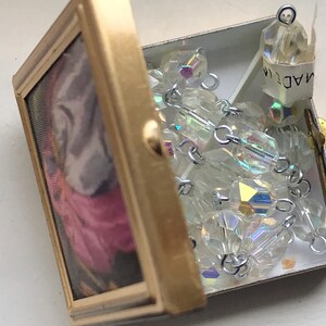 Puede incluir: Una caja de metal de color dorado con un dise&ntilde;o floral en el interior. La caja est&aacute; abierta y contiene una hebra de cuentas de cristal iridiscente con conectores de metal plateado. La etiqueta en las cuentas dice "MADE IN".