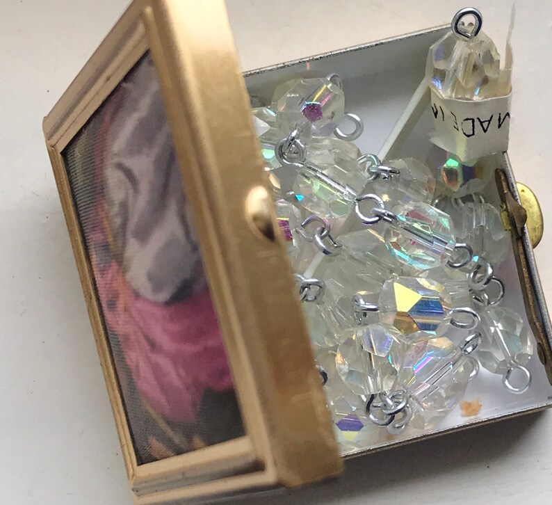 Puede incluir: Una caja de metal de color dorado con un dise&ntilde;o floral en el interior. La caja est&aacute; abierta y contiene una hebra de cuentas de cristal iridiscente con conectores de metal plateado. La etiqueta en las cuentas dice "MADE IN".