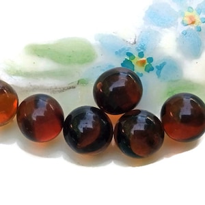 May include: Six brown glass marbles with a mottled, marbled effect. The marbles are arranged in a semi-circle on a white surface with a floral design.