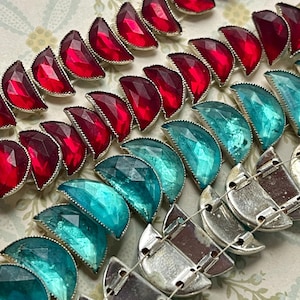 May include: A close-up of three rows of half-moon shaped glass beads. The top row is red, the middle row is teal, and the bottom row is silver.
