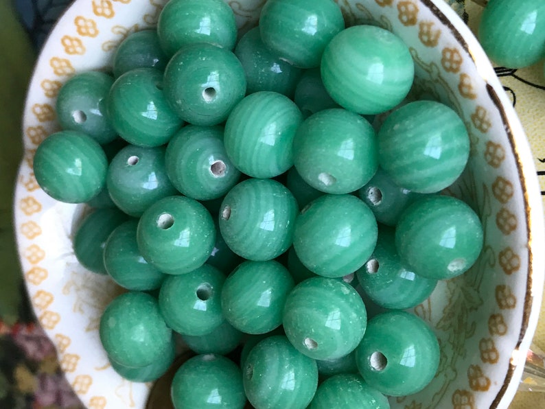 May include: A bowl filled with smooth, round, green jade beads. The beads have a subtle swirl pattern and a hole in the center.