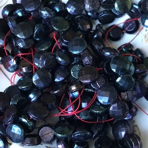 Puede incluir: Una hebra de cuentas de vidrio azul oscuro iridiscente con un hilo rojo que las atraviesa. Las cuentas son redondas y tienen una superficie facetada.