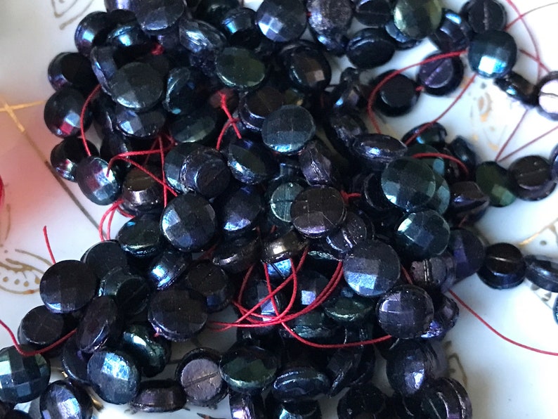 Puede incluir: Una hebra de cuentas de vidrio azul oscuro iridiscente con un hilo rojo que las atraviesa. Las cuentas son redondas y tienen una superficie facetada.