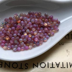 K&ouml;nnte beinhalten: Nahaufnahme eines wei&szlig;en L&ouml;ffels, der mit schimmernden rosa und violetten Perlen gef&uuml;llt ist. Die Perlen sind rund und haben einen leichten Glanz.
