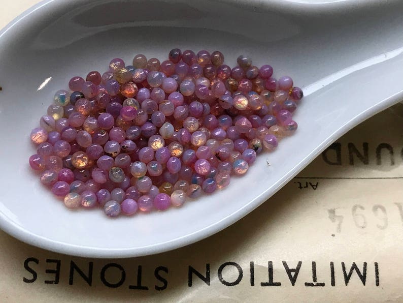 K&ouml;nnte beinhalten: Nahaufnahme eines wei&szlig;en L&ouml;ffels, der mit schimmernden rosa und violetten Perlen gef&uuml;llt ist. Die Perlen sind rund und haben einen leichten Glanz.