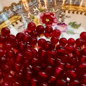 May include: A bowl filled with deep red, round, glass beads. The beads are smooth and shiny, and they are arranged in a random pattern.