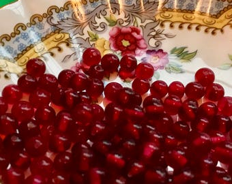 May include: A bowl filled with deep red, round, glass beads. The beads are smooth and shiny, and they are arranged in a random pattern.