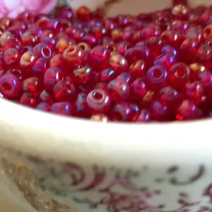 May include: A close-up of a bowl filled with small, iridescent red beads. The beads have a slightly metallic sheen and are arranged in a random pattern.