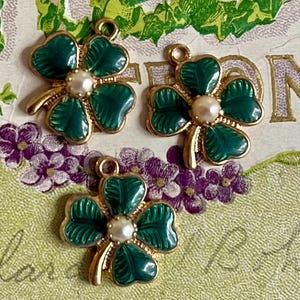 Puede incluir: Tres colgantes dorados con forma de tr&eacute;bol de cuatro hojas, con hojas de esmalte verde y una perla central. Los colgantes tienen un lazo para sujetarlos a la joyer&iacute;a. El fondo presenta un dise&ntilde;o floral con flores moradas y follaje verde.