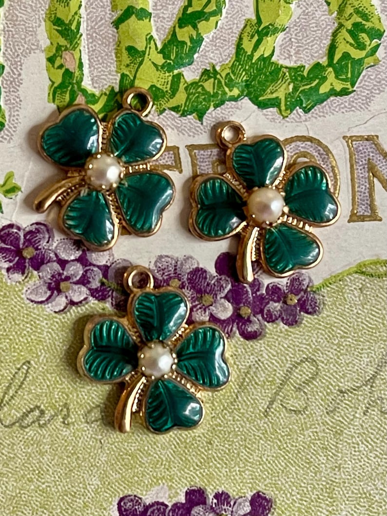 Puede incluir: Tres colgantes dorados con forma de tr&eacute;bol de cuatro hojas, con hojas de esmalte verde y una perla central. Los colgantes tienen un lazo para sujetarlos a la joyer&iacute;a. El fondo presenta un dise&ntilde;o floral con flores moradas y follaje verde.
