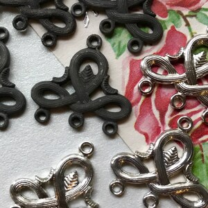 May include: A close-up of eight metal decorative pieces. Four are silver and four are black. Each piece is shaped like a stylized heart with three loops at the bottom. The pieces are arranged in a random pattern.