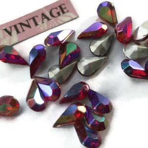 May include: A collection of vintage iridescent teardrop-shaped rhinestones in a deep red color. The rhinestones have a faceted design and are scattered on a white surface. The word "Vintage" is printed on a pink and white label in the background.