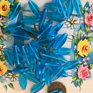 May include: A pile of turquoise blue glass beads in the shape of long oval leaves. The beads are scattered on a white surface with a floral pattern.