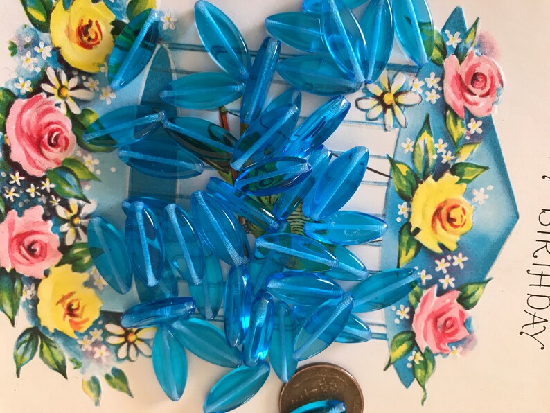 May include: A pile of turquoise blue glass beads in the shape of long oval leaves. The beads are scattered on a white surface with a floral pattern.