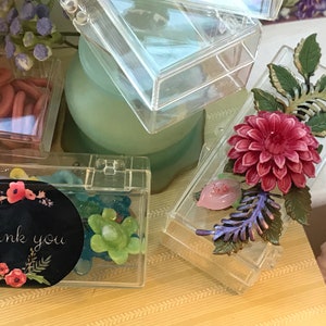 May include: Four clear plastic boxes with various small items inside. The boxes are on a yellow and white patterned surface. One box has a black and white sticker that says "thank you" with small red flowers. Another box has a pink flower with green leaves.