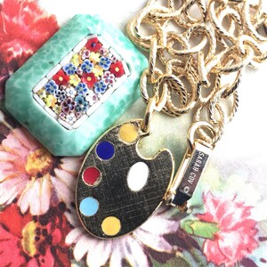 K&ouml;nnte beinhalten: Goldkette mit einem gr&uuml;nen und wei&szlig;en Mosaik-Anh&auml;nger und einem goldenen, bemalten K&uuml;nstler-Palette-Anh&auml;nger. Der Palette-Anh&auml;nger hat rote, blaue, gelbe und wei&szlig;e Farben. Die Kette hat einen Verschluss mit der Aufschrift "SARAH COV."