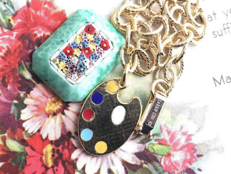 K&ouml;nnte beinhalten: Goldkette mit einem gr&uuml;nen und wei&szlig;en Mosaik-Anh&auml;nger und einem goldenen, bemalten K&uuml;nstler-Palette-Anh&auml;nger. Der Palette-Anh&auml;nger hat rote, blaue, gelbe und wei&szlig;e Farben. Die Kette hat einen Verschluss mit der Aufschrift "SARAH COV."