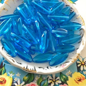 May include: A bowl filled with turquoise blue glass beads. The beads are oval shaped and have a smooth, glossy finish.
